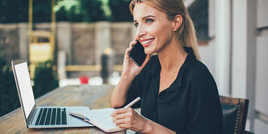 Happy female student receiving positive cellphone call sitting outdoors with laptop device and education textbook for e learning, prosperous freelancer communicate via smartphone and smiling